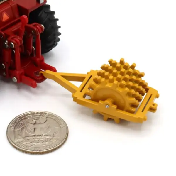 A small yellow toy agricultural roller attached to a red toy tractor next to a coin.